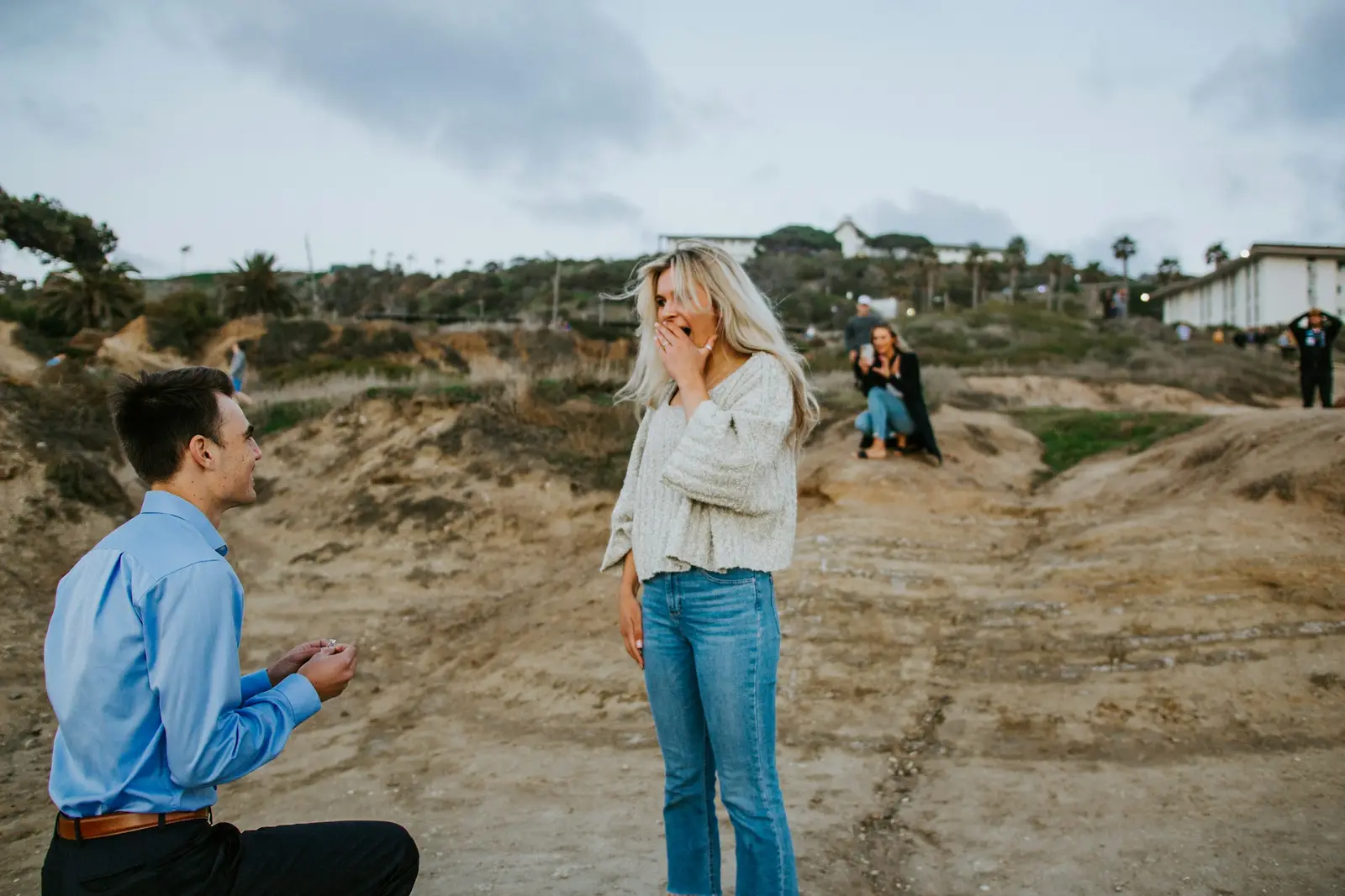 A person on one knee proposing on a quiet park path as their partner covers their mouth in surprise